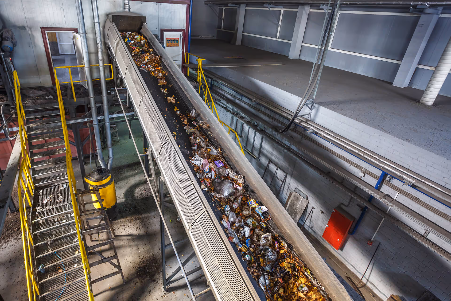Overhead view of a long conveyor belt carrying a mix of trash and waste material inside a recycling or waste sorting facility, next to yellow stairs.